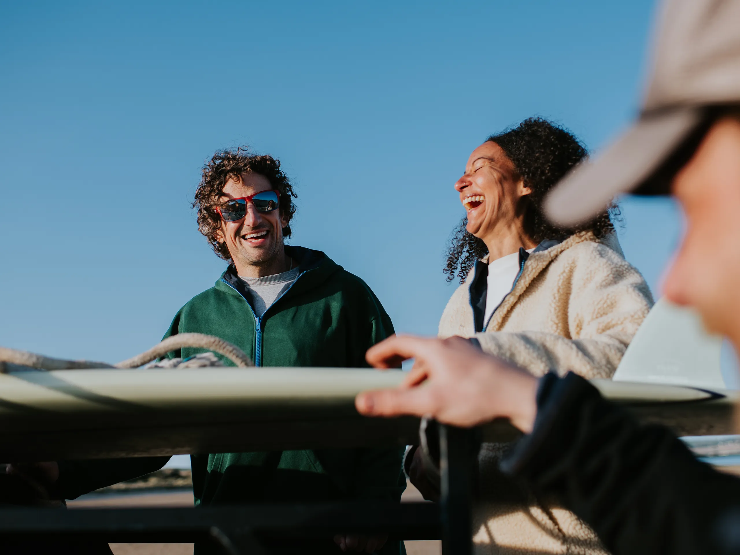 Dos personas riendo juntas en la playa junto a una tabla de surf.