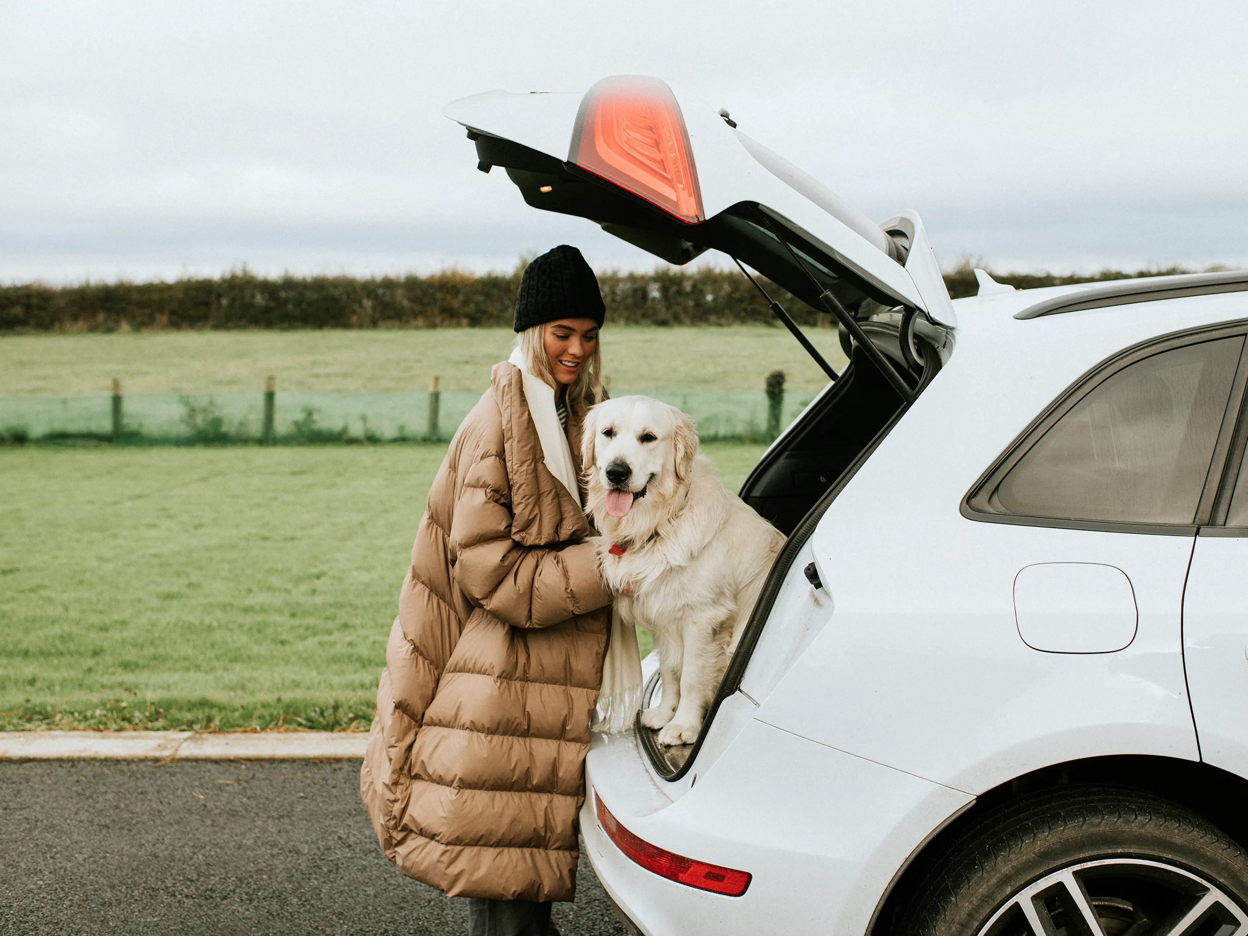 Una mujer con un golden retriever está de pie junto a un coche con el maletero abierto.