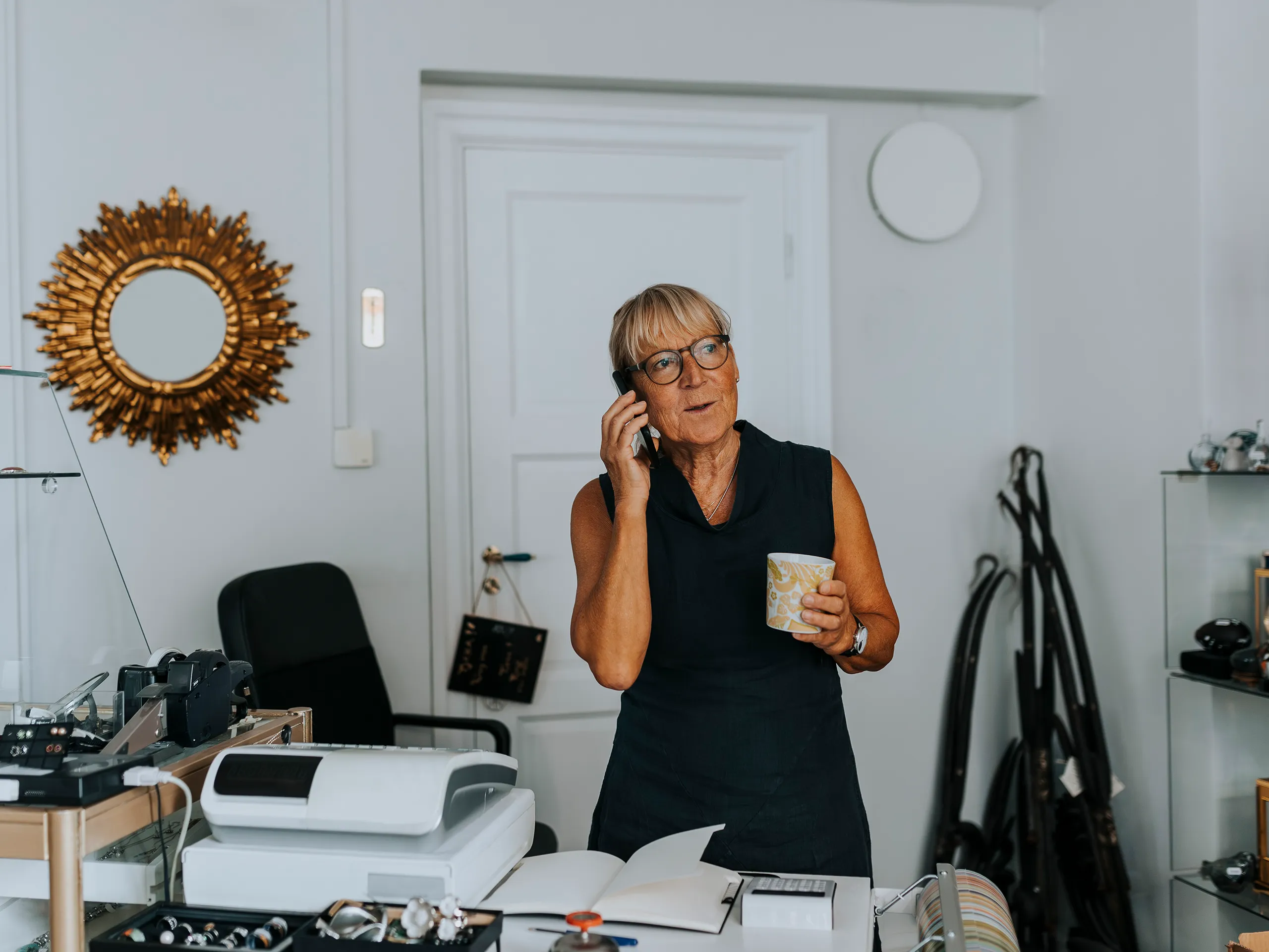 Woman with glasses on phone in shop, holding a cup.