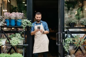 Hombre blanco dueño de floristeria, en la puerta de su negocio mirando el móvil